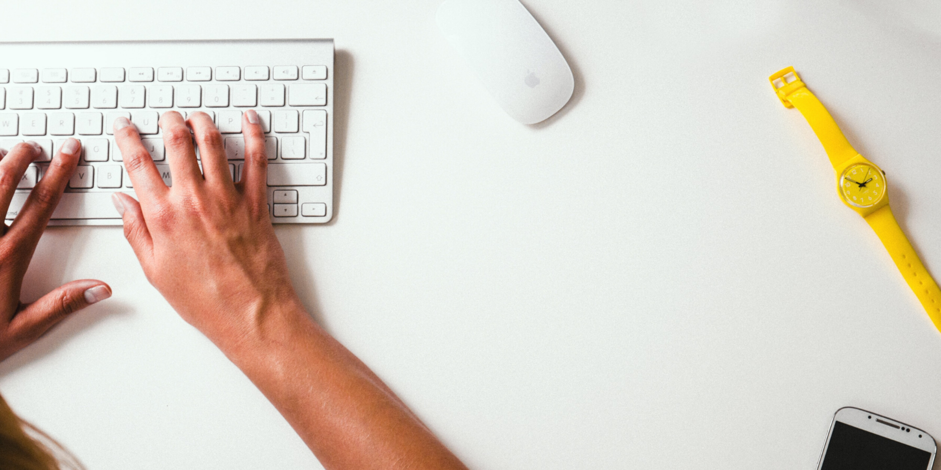 Hands typing on a keyboard on a white desk with mouse, watch, and phone on top